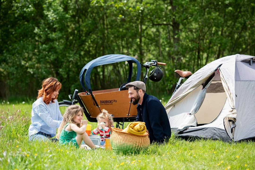 Ein Bild von einer Familie die vor einem Babboe City Mountain sitzt und Picnict, neben dran steht ein Zelt.