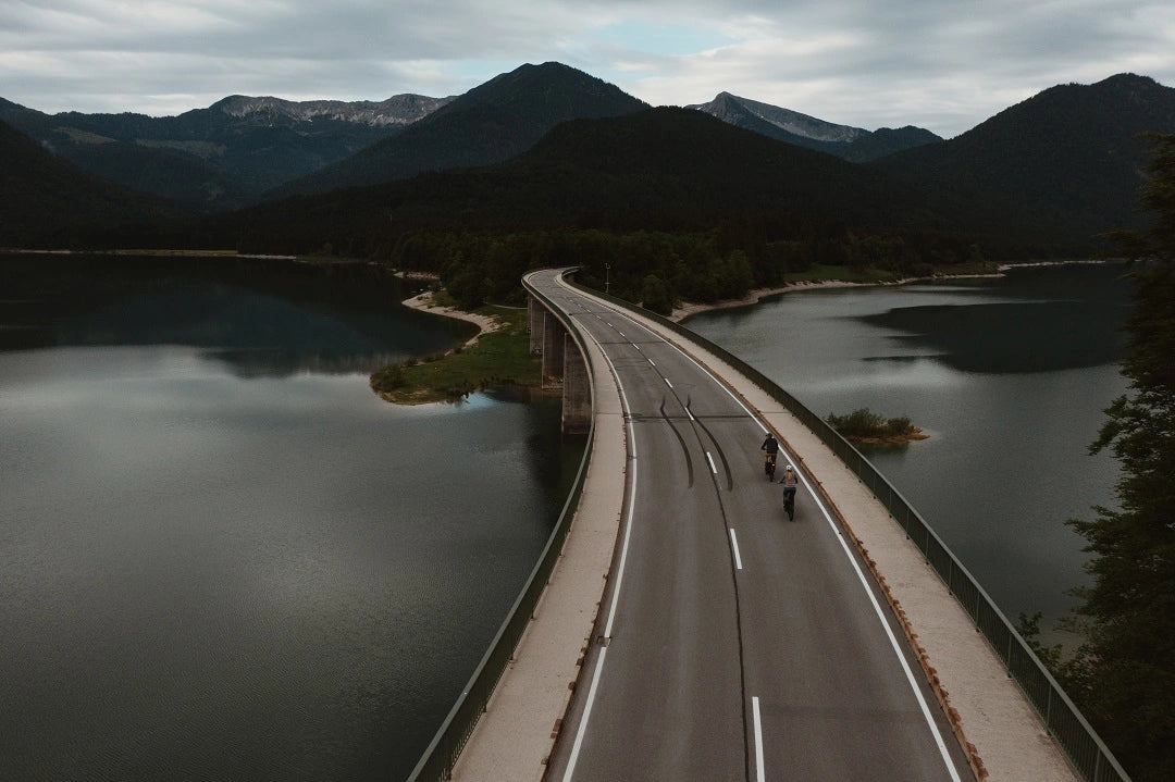 Zwei Radfahrer fahren über eine Brücke die über einen See verläuft und zwischen vielen Bergen mündet